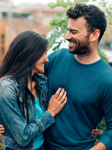 A married couple standing close together outdoors, smiling warmly at each other